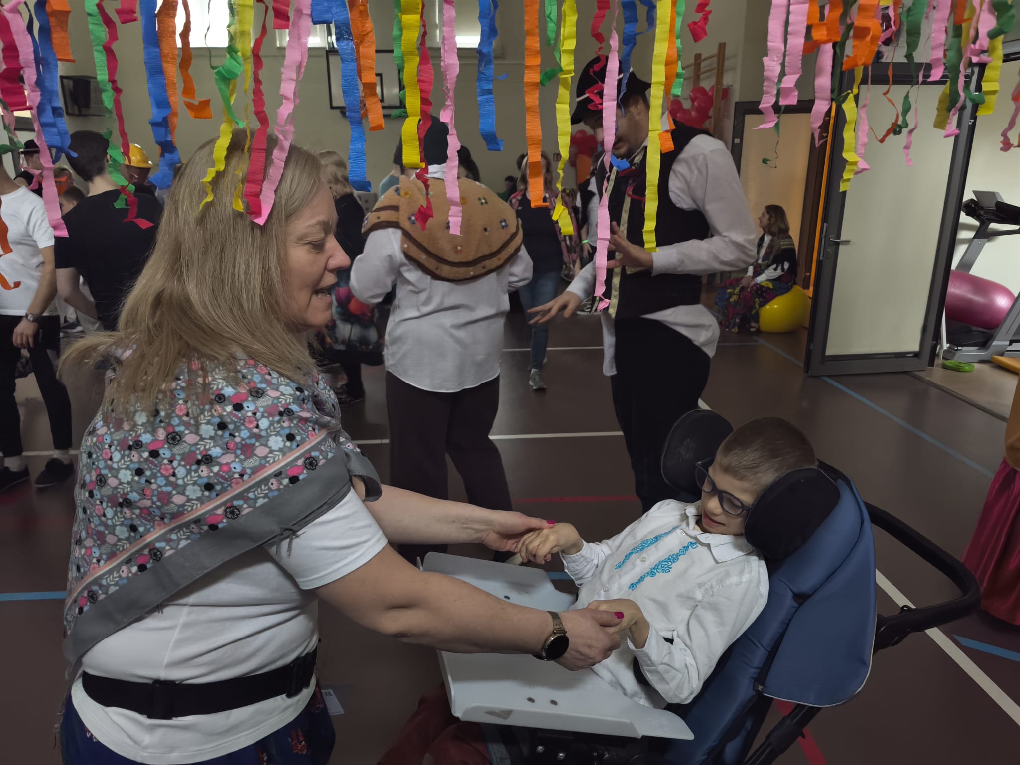 Kobieta tańczy z chłopcem siedzącym na wózku. W tle znajdują się tańczący uczniowie. 