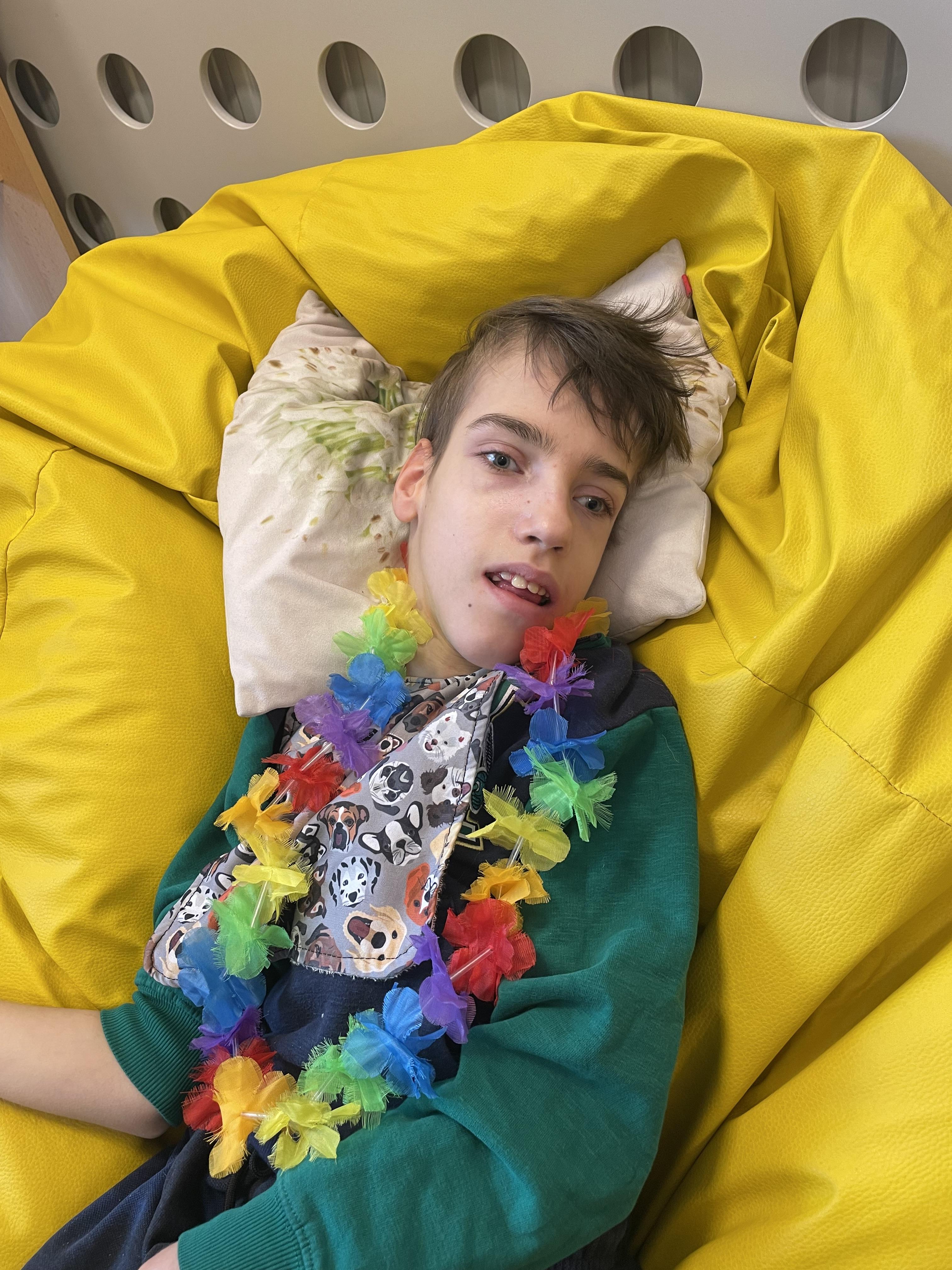 Chłopiec leży na żółtej pufie. Na szyi ma naszyjnik hawajski- kolorowe kwiaty. 