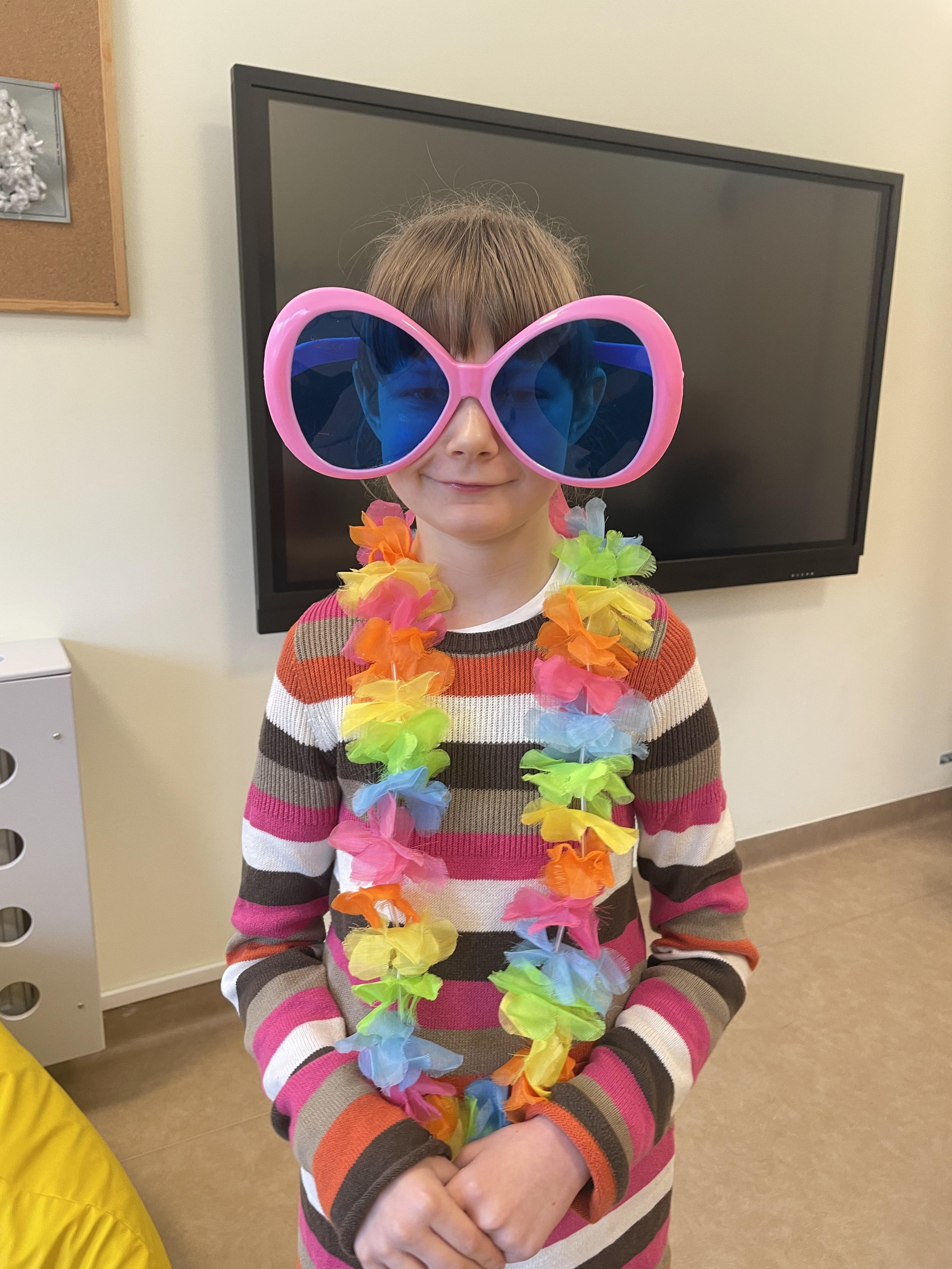 Uśmiechnięta dziewczynka,  ubrana w sweter w kolorowe paski, stoi przed ekranem tablicy multimedialnej. Na nosie ma duże okulary, które mają różowe oprawki i niebieskie szkła. Na szyi ma założony hawajski naszyjnik.