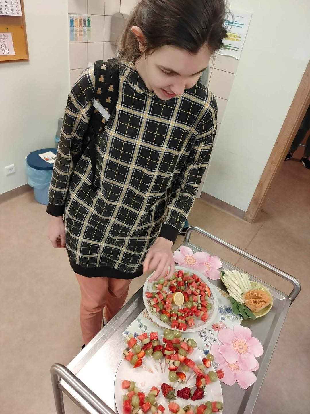 Uczennica stoi obok wózka kelnerskiego, na którym znajdują się dwa talerze z owocowymi koreczkami z winogrona, kiwi, truskawki i arbuza. 