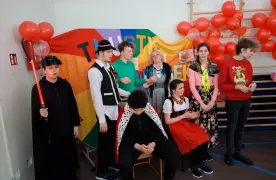 Na zdjęciu znajduje się 5 uczniów, 2 uczennice i nauczycielka. Za nimi znajduje się kolorowej tło z napisem tłusty czwartek i balony w kolorze czerwonym. 