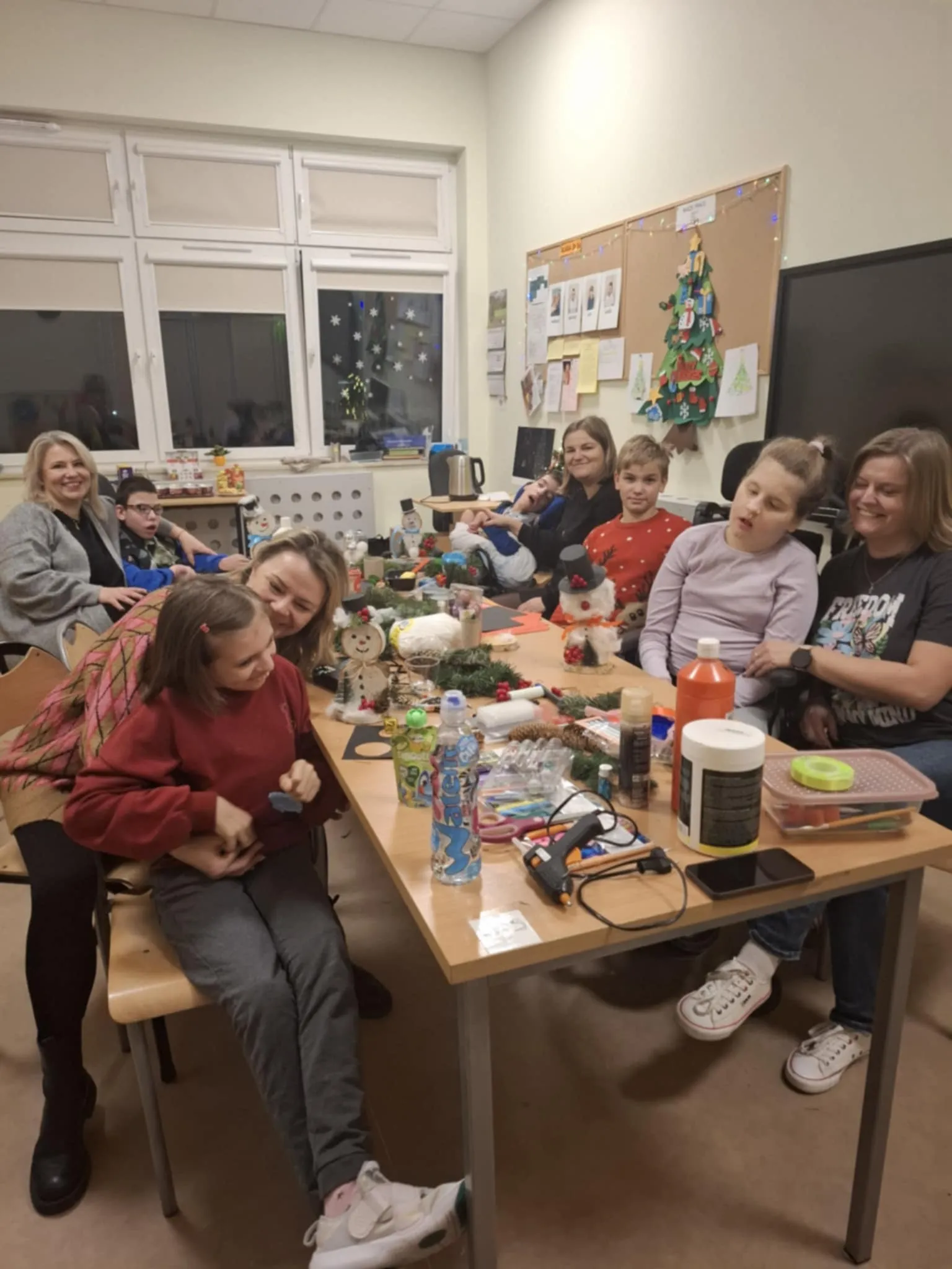 Uczestnicy warsztatów siedzą przy stole zastawionym różnymi przyborami plastycznymi. Wszyscy uczestnicy uśmiechają się i pozują do zdjęcia.