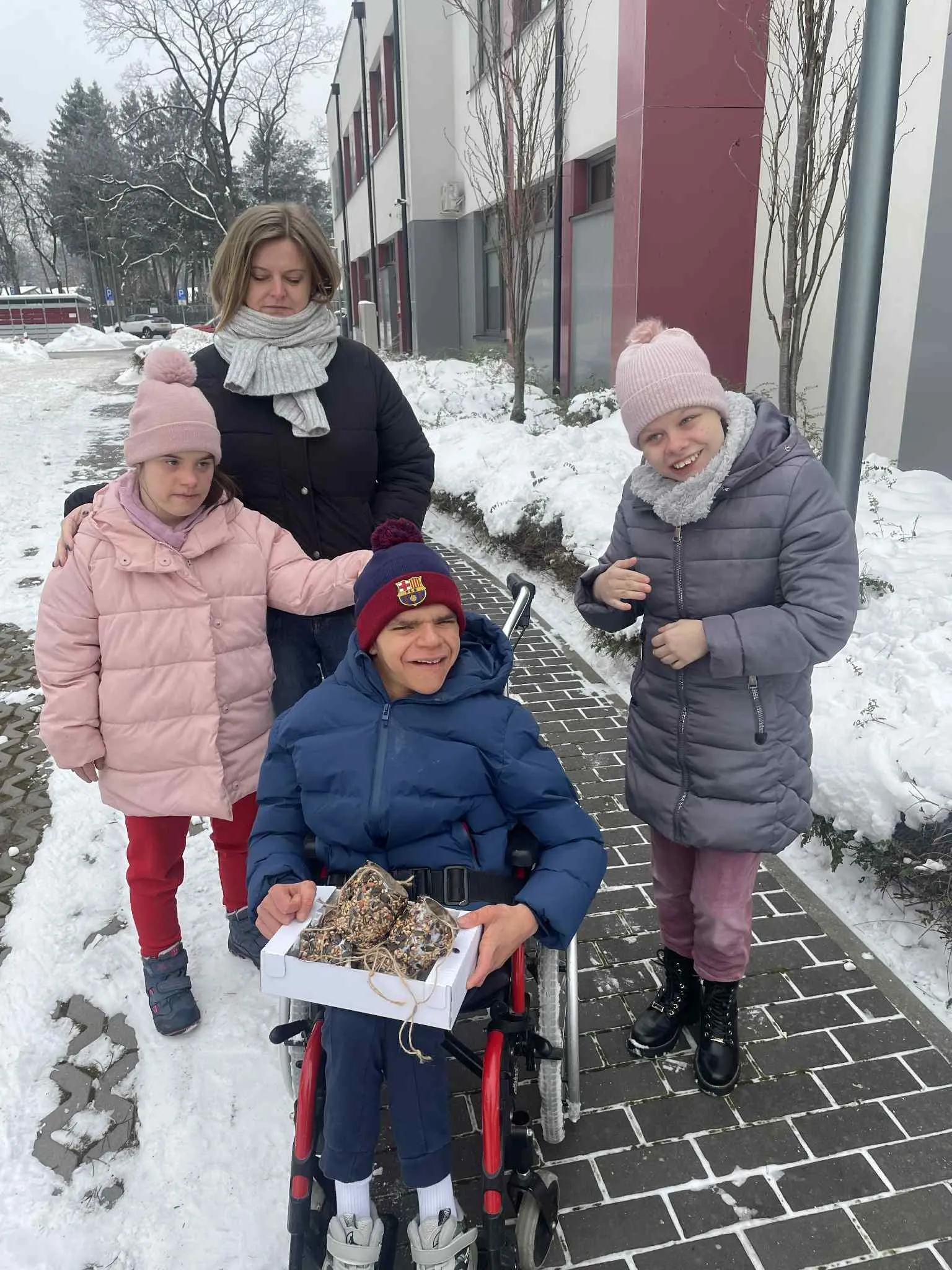Na boisku szkolnym jest chłopiec na wózku inwalidzkim wokół niego stoją dwie dziewczyny i nauczycielka. Chłopiec na kolanach trzyma pudełko, w  którym znajdują się proste karmniki wykonane z tekturowej rolki, sznurka, smalcu i ziaren.