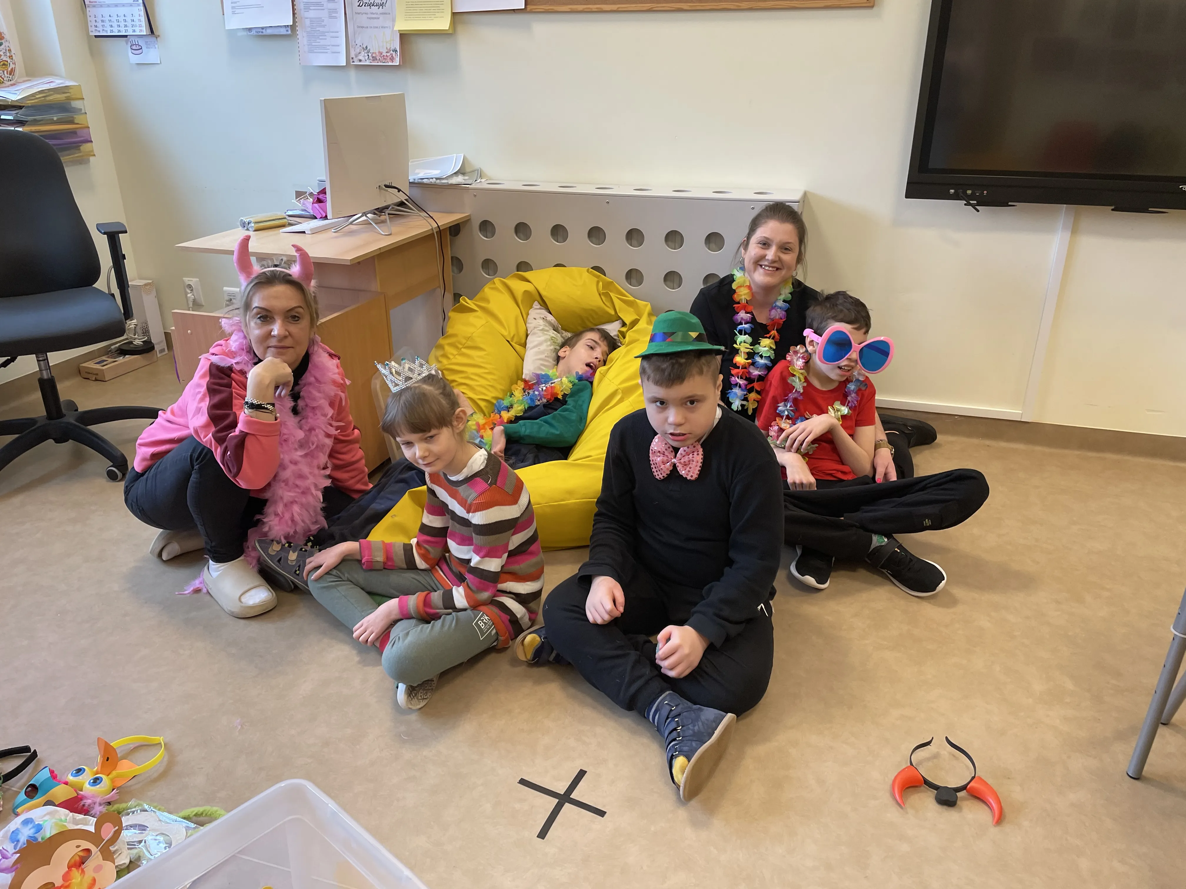 Na zdjęciu są dwie kobiety, jedna dziewczynka i trzech chłopców. Chłopiec siedzący na żółtej pufie, kobieta siedząca obok niego i chłopiec siedzący obok kobiety mają na szyi ubrane naszyjniki hawajskie. Przed uczniem siedzącym na pufie znajduje się kobieta, która ubrana jest w różowy szal z piórek a na głowie ma opaskę różowe rogi. Jest również dziewczynka, która ma koronę na głowie. Obok dziewczynki siedzi chłopiec w zielonym meloniku na głowie i różową muszką pod brodą. 