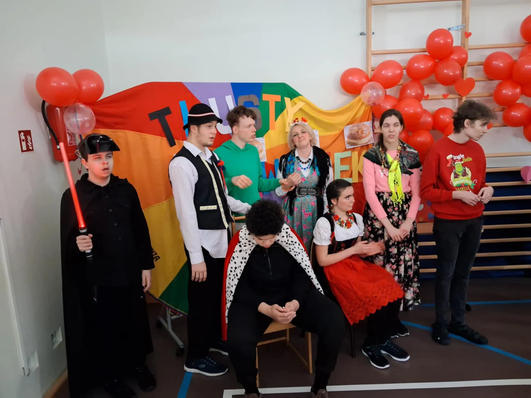 Na zdjęciu znajduje się 5 uczniów, 2 uczennice i nauczycielka. Za nimi znajduje się kolorowej tło z napisem tłusty czwartek i balony w kolorze czerwonym. 