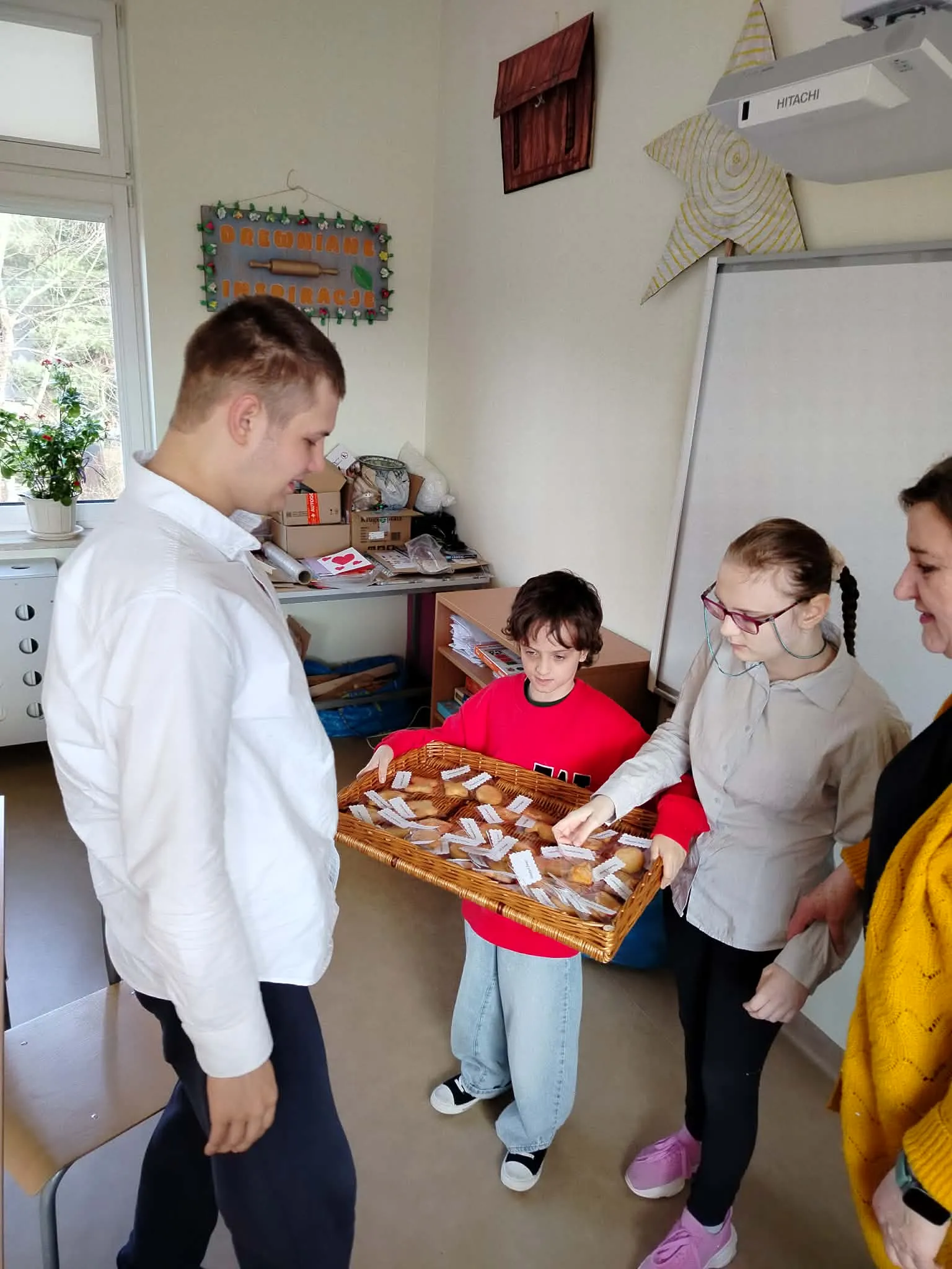 Chłopiec w czerwonej bluzie trzyma wiklinową tacę z ciasteczkami. Przed  nim stoi młody, uśmiechnięty mężczyzna, który oczekuje na otrzymanie słodkiego upominku od dziewczynki w okularach, stojącej obok chłopca w czerwonej bluzie.  