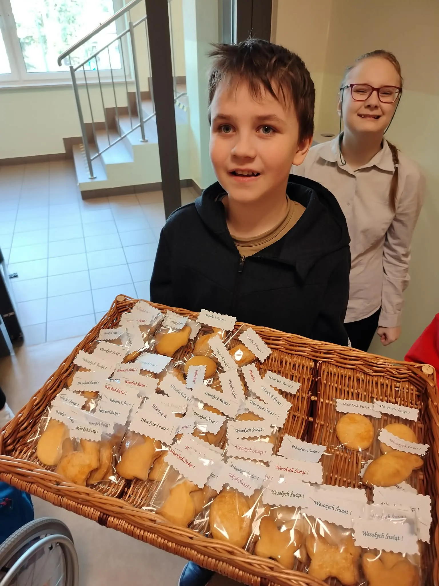 Chłopiec trzyma wiklinową tacę na której ułożone są zapakowane ciasteczka w kształcie jajka, krolika, marchewki, motyla, z przypiętą kartką Wesołych Świąt. Za chłopców stoi uśmiechnięta dziewczynka w okularach. 