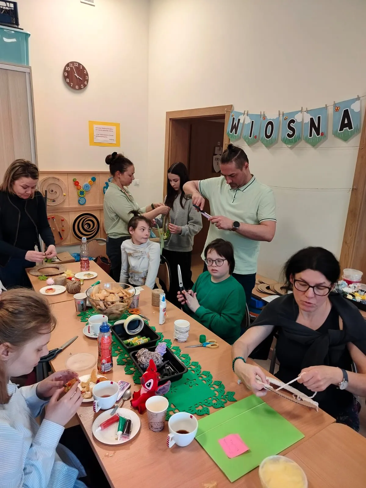 Na zdjęciu znajdują się dziewczynki, kobiety i jeden mężczyzna. 3 dziewczynki i dwie kobiety siedzą przy stole, wycinają, nawlekają sznurek na kartonowy wianek, dekorują ciasteczka. Na stole znajdują się różne materiały potrzebne do wykonania wianków wiosenno wielkanocnych.  Dwie stojące kobiety mocują wstążkę na wianku a  mężczyzna tnie materiał. 