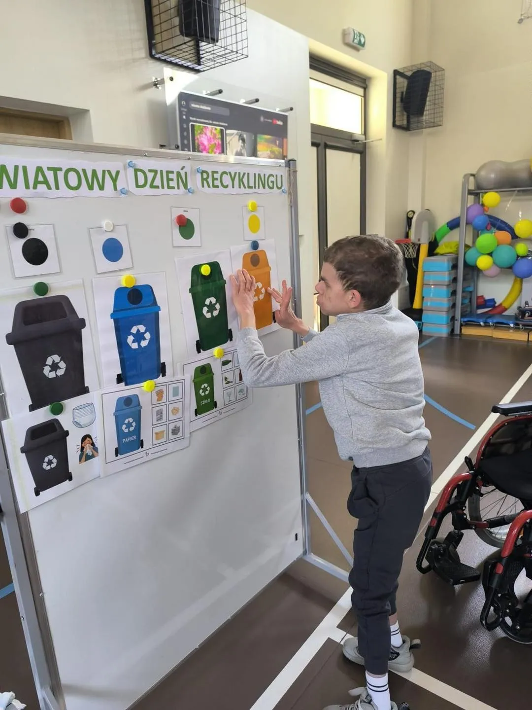 Chłopiec stoi przy tablicy, na której znajduje się napis światowy dzień recyklingu. Pod napisem  na wydrukowanych kartkach są 3 kosze  na śmieci w kolorach: czarny, niebieski, zielony, 4 w kolorze żółtym przyczepia chłopiec. 