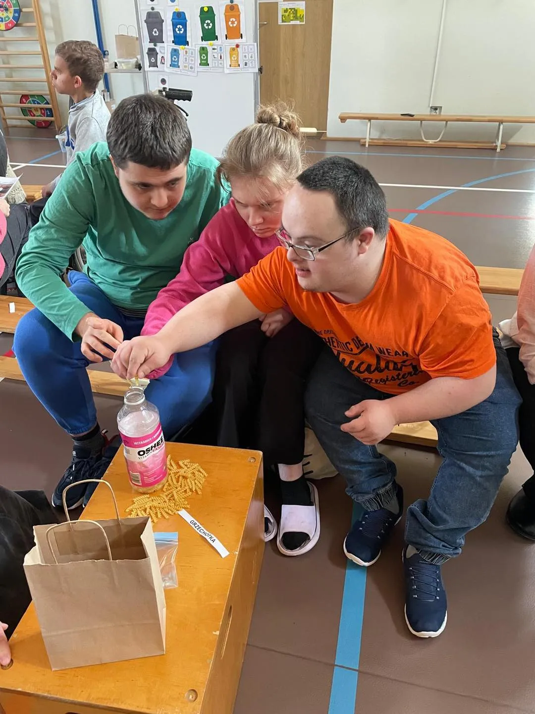 Jedna uczennica i dwóch uczniów siedzą na ławce,  wkładają makaron do butelki plastikowej.  Ich zadaniem jest przygotować grzechotkę z materiałów recyklingowych. 