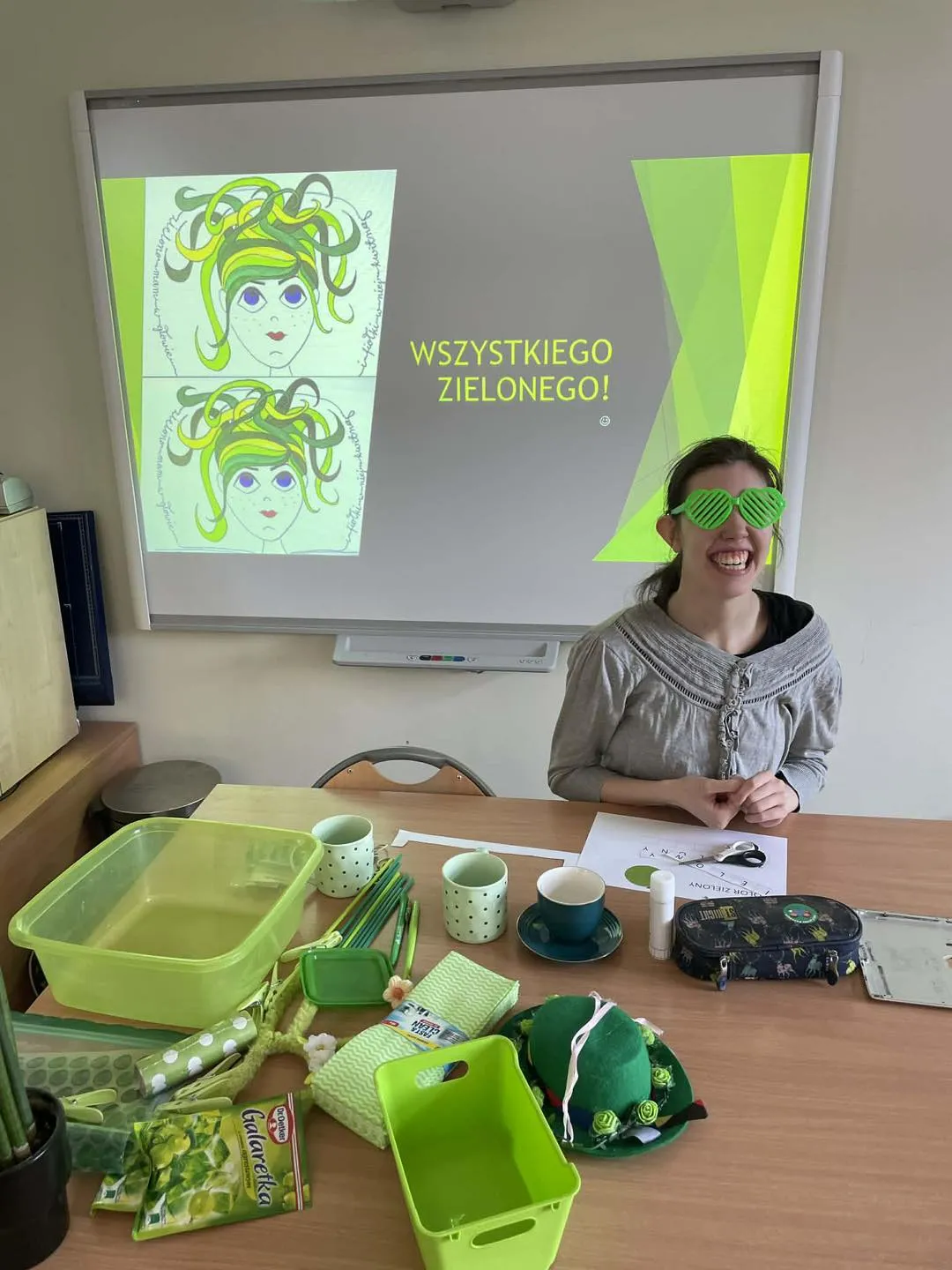 Uczennica siedzi przy stole, ma założone zielone okulary, uśmiecha się. Na stole znajdują się różne zielone przedmioty: pudełka, spinacze, kubki, miska, galaretki. Za uczennicą jest tablica multimedialna na której wyświetlony jest napis wszystkiego zielonego i obok zdjęcie kobiety z zielonymi włosami.
