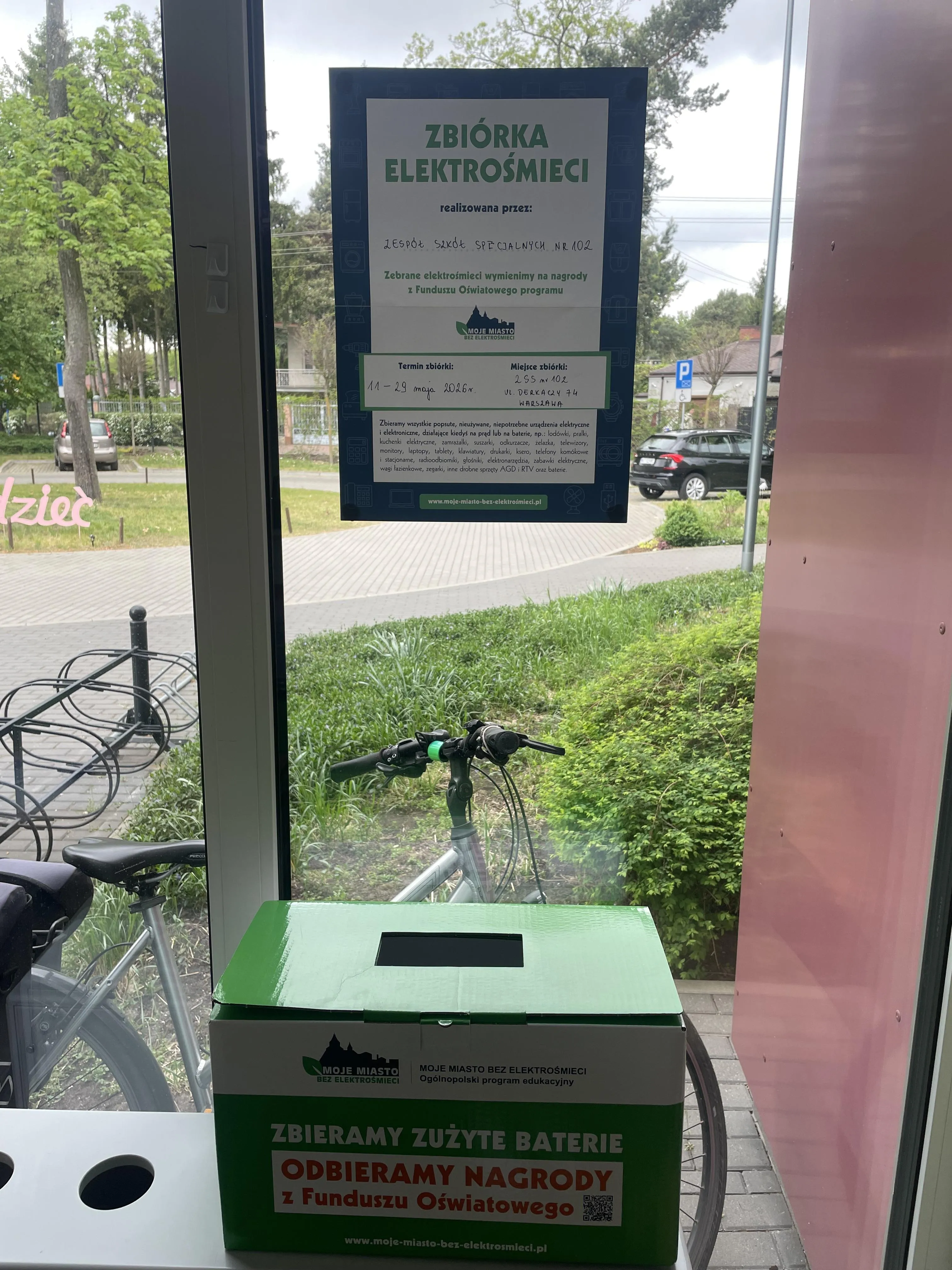 Zdjęcie przedstawia plakat informujący o zbiórce elektrośmieci. Plakat wisi na oknie pod nim znajduje się zielone pudełko z otworem na baterie. Na plakacie znajduje się informacja, że zbiórka elektrośmieci organizowana jest przez Zespół Szkół Specjalnych nr 102 w Warszawie w dniach od 11 do 29 maja 2026r. 